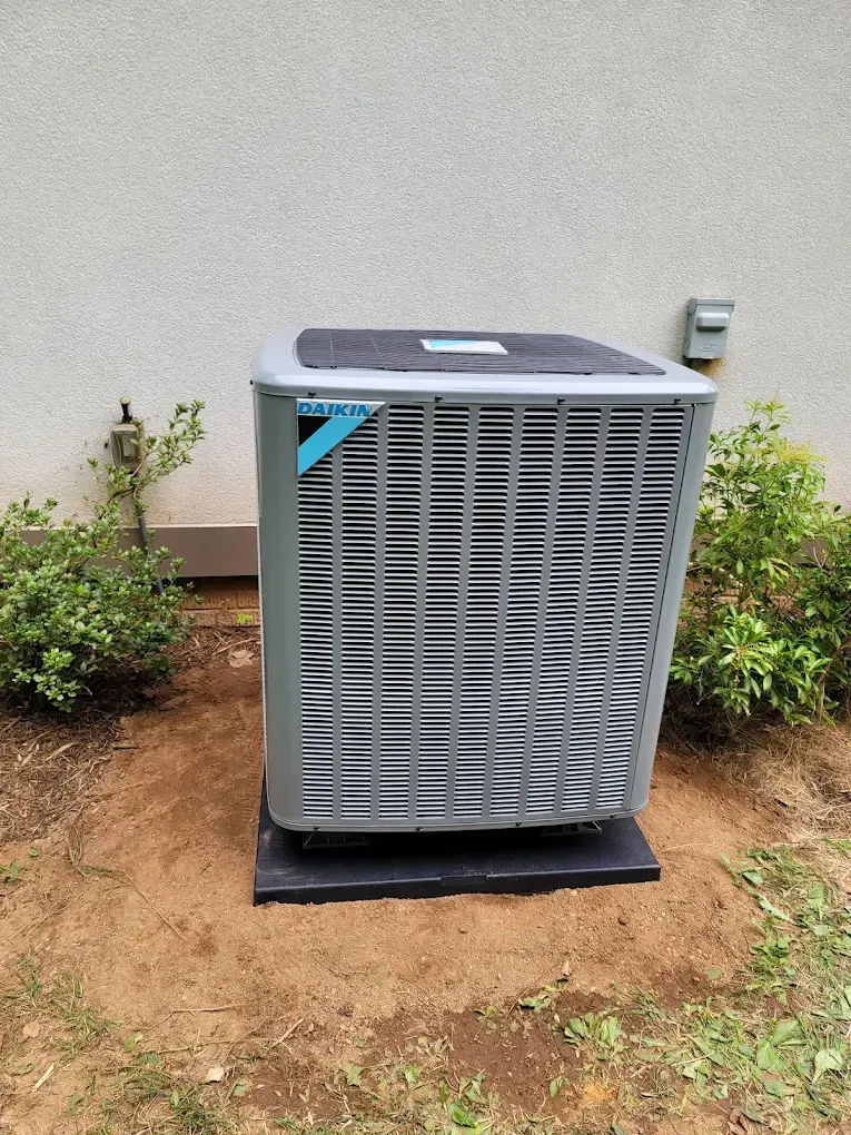 Outdoor HVAC unit on a black pad beside shrubs against a beige wall.