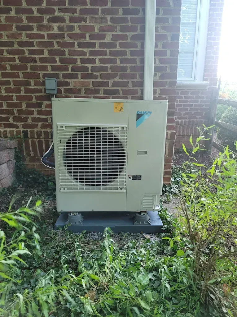 Outdoor HVAC unit beside a brick wall, surrounded by overgrown grass and plants