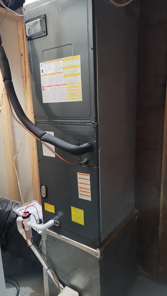 Gray HVAC furnace unit with attached black ducting and labels in a utility room.