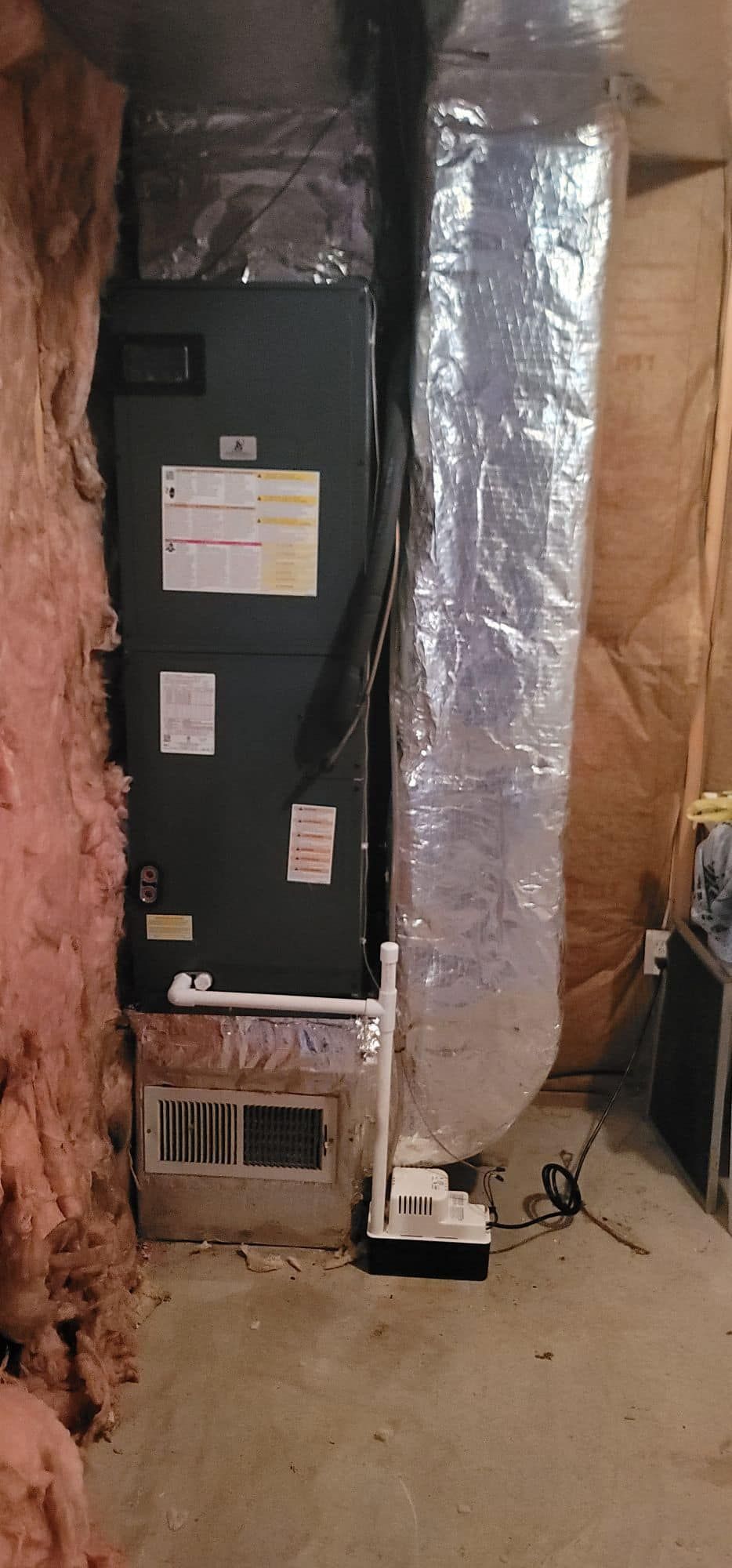 Tall black HVAC unit with silver ducting in an unfinished basement next to exposed insulation and concrete floor