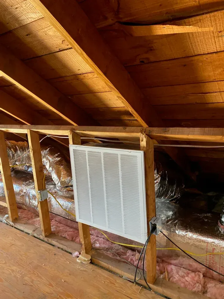 Attic with exposed wooden rafters, insulation, and a white wall vent or filter panel.