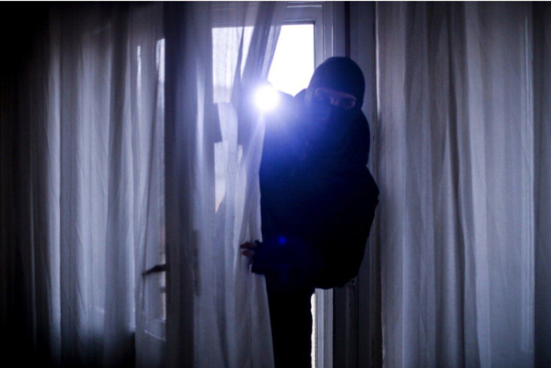 A masked figure, silhouetted in a dark room, peers through a window with bright light.