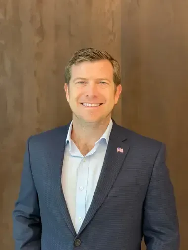 Man in blue blazer smiles in front of a wooden wall.