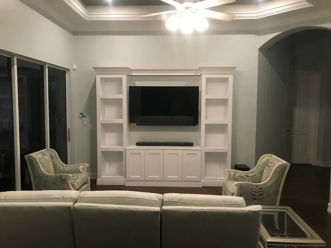 Living room with white entertainment center, TV, gray sofa, and two patterned armchairs under a ceiling fan.