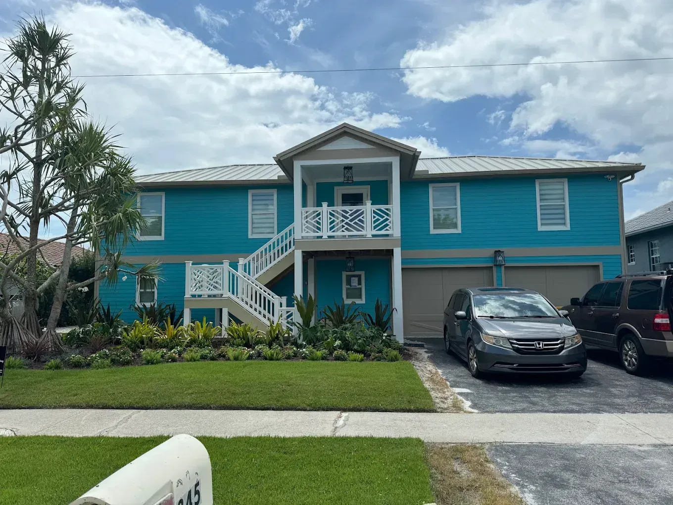 Turquoise two-story house with white trim, front porch, garage, and parked cars under a cloudy sky