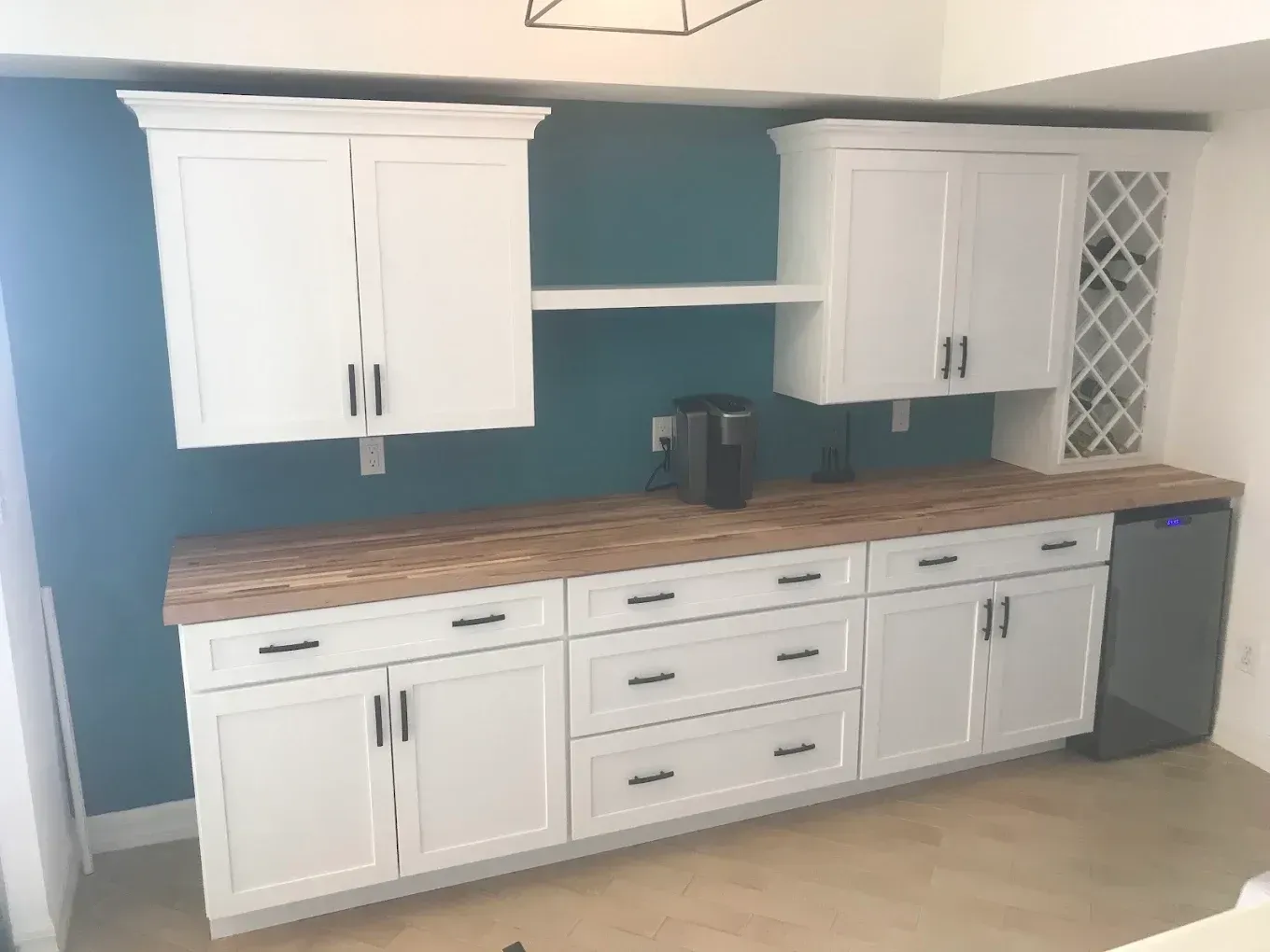 White kitchen cabinets with wooden countertops against a teal wall, featuring a wine rack on the right.