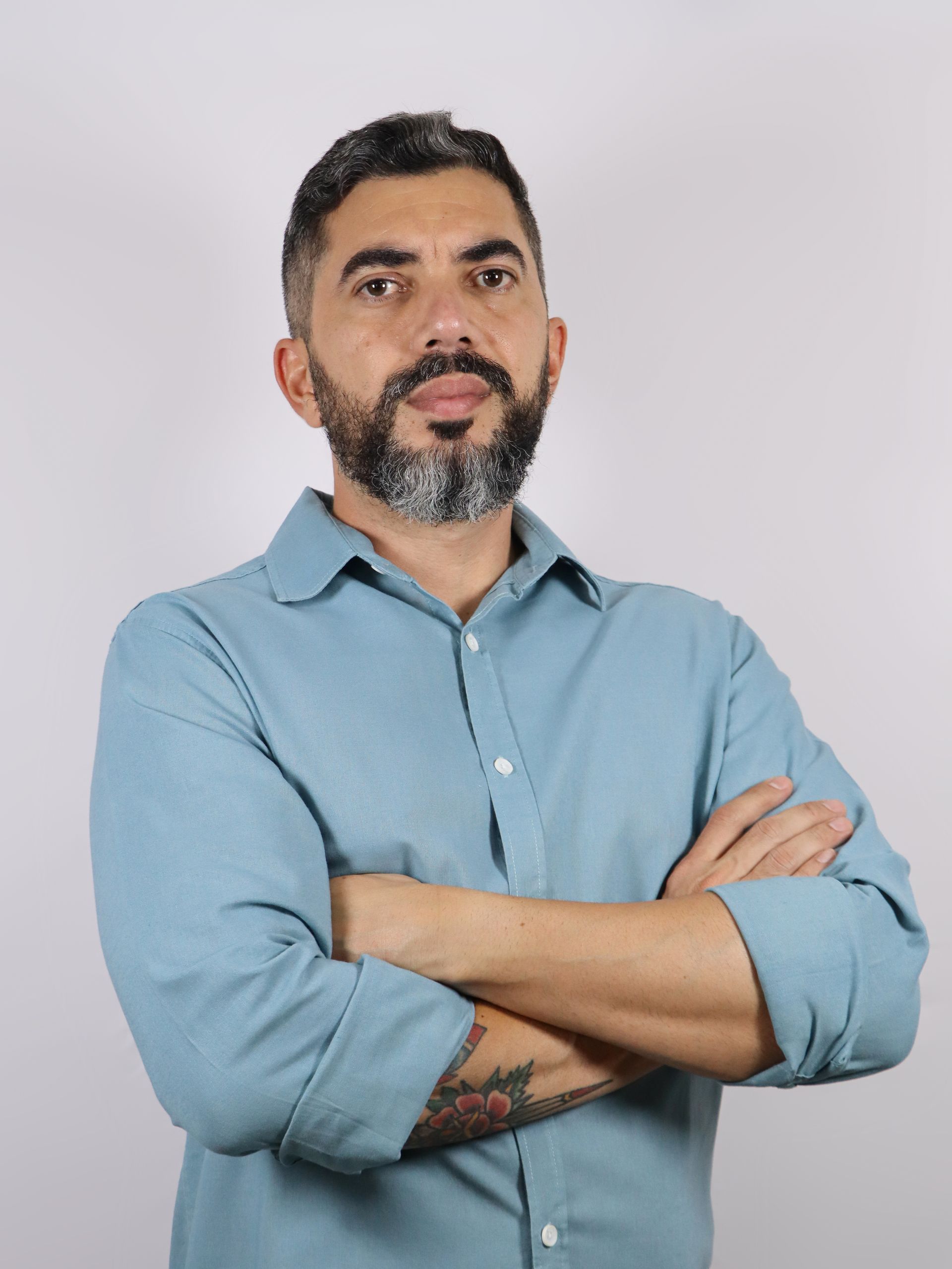 Um homem com barba está de pé com os braços cruzados.