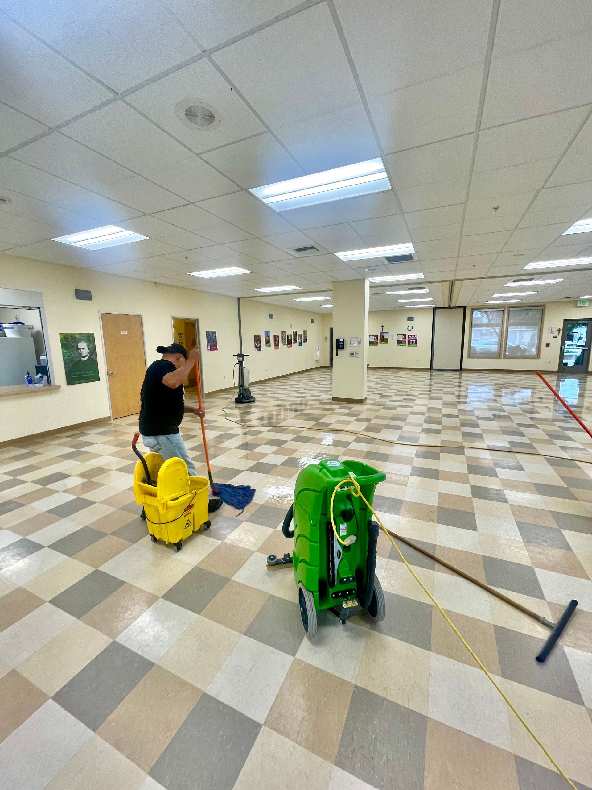 Person mopping a large tiled floor with a yellow bucket, green cart, and a squeegee in a brightly lit room.