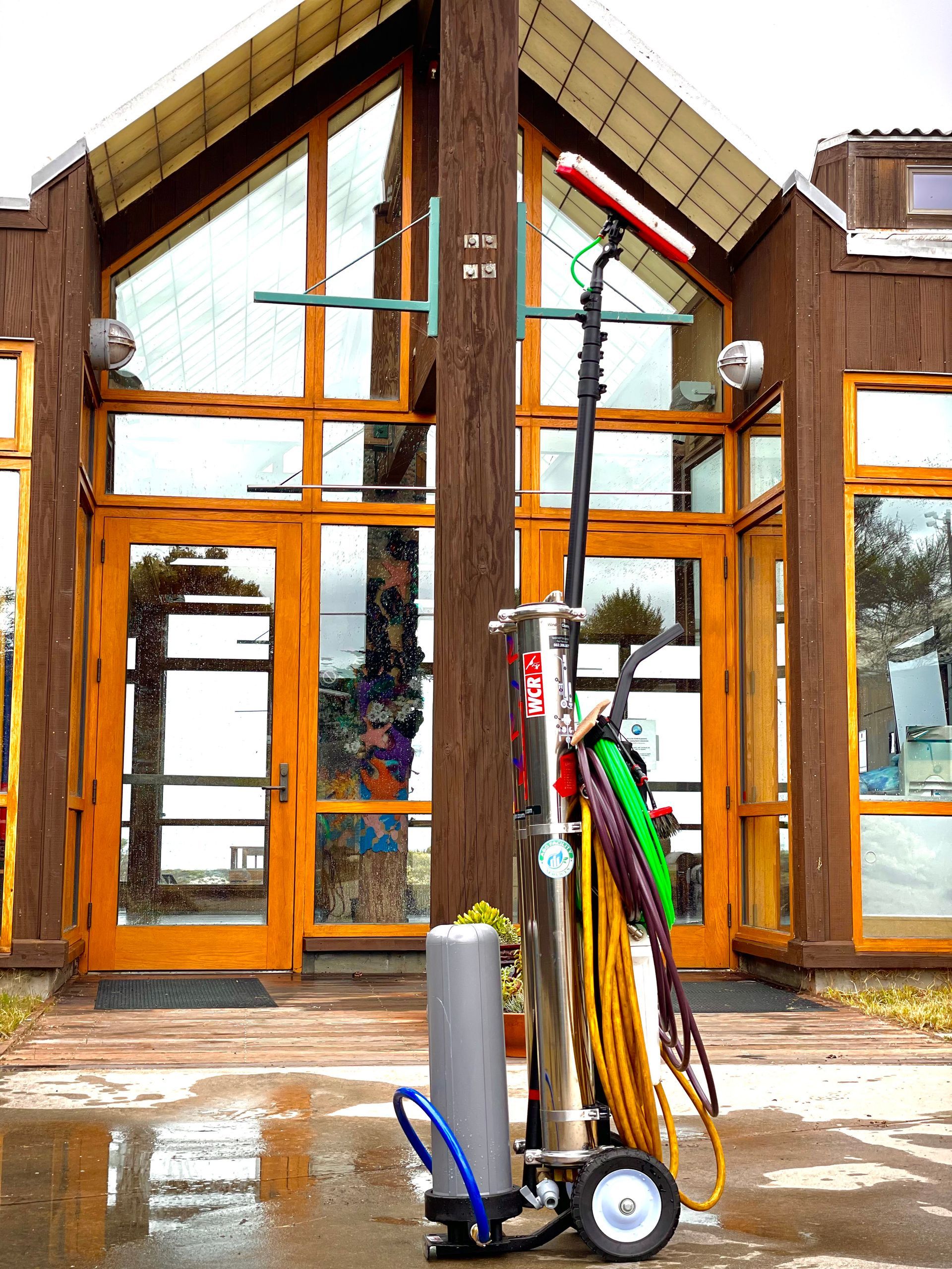 A portable water-fed window cleaning system stands on a wet pavement in front of a wood-framed glass building.