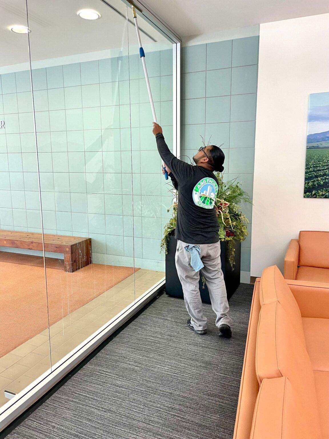 A person wearing a black long-sleeved shirt and gray pants uses a long-handled squeegee to clean a tall glass wall.