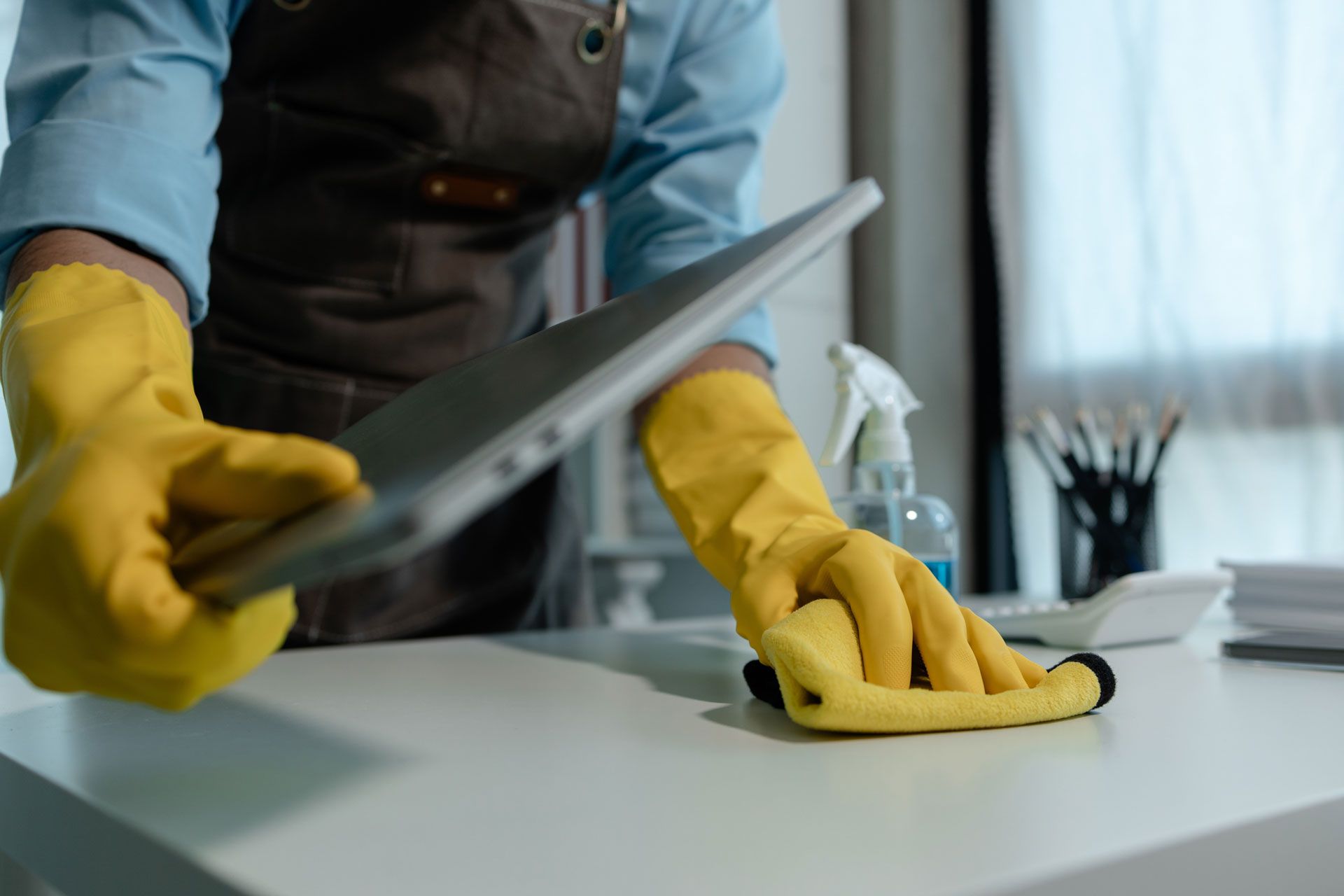 A person wearing yellow rubber gloves and an apron wipes a white desk while holding a tablet.