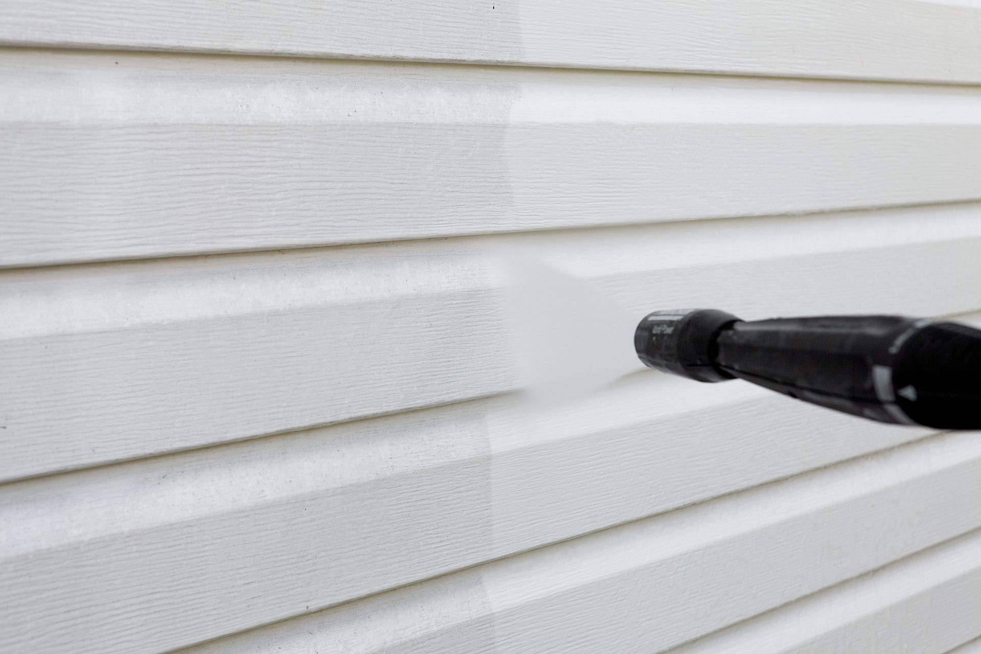 A person is cleaning a white siding with a high pressure washer.