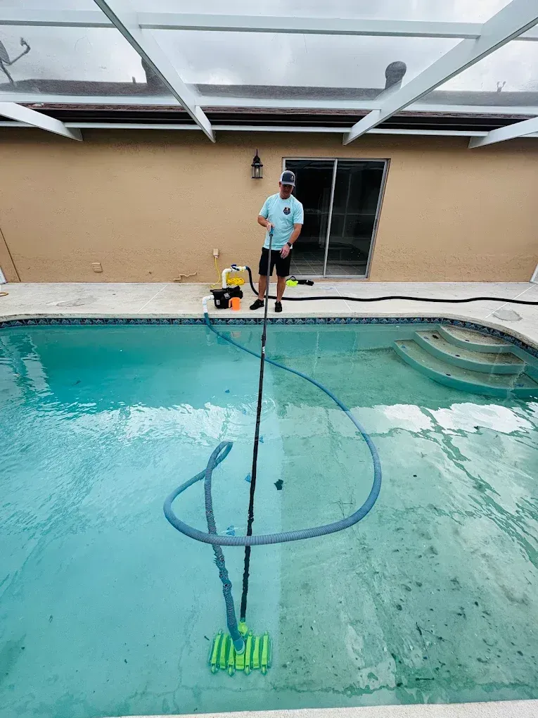 Person cleaning a swimming pool with a long-handled vacuum, cloudy sky overhead.