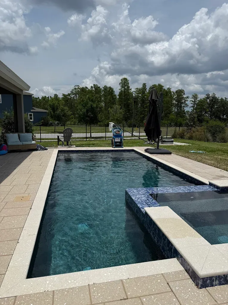 Pool with dark water, stone patio, lounge area, trees in the distance, and a partially cloudy sky.