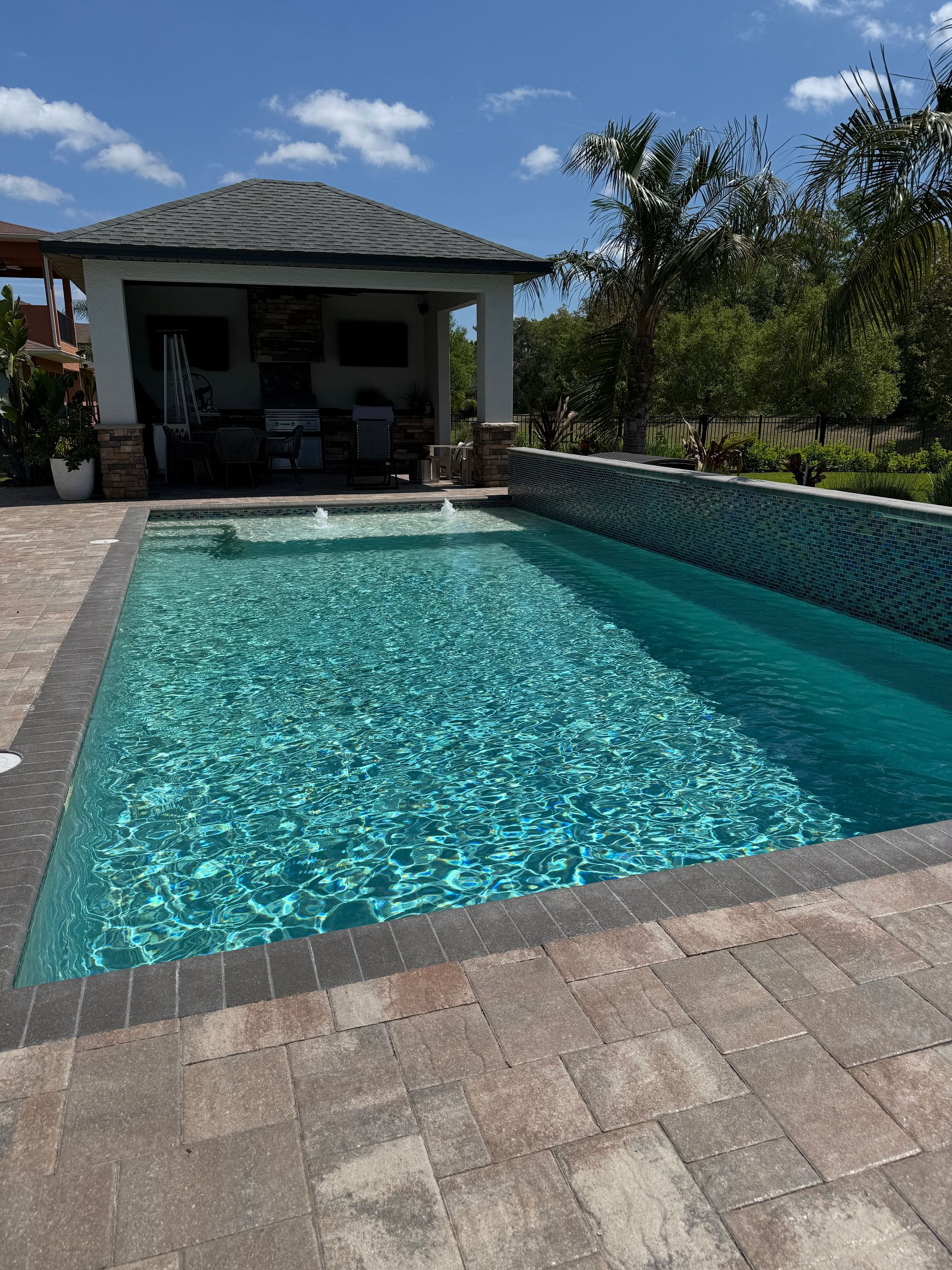 Rectangular pool with turquoise water, a brick patio, and a covered patio area under a blue sky.