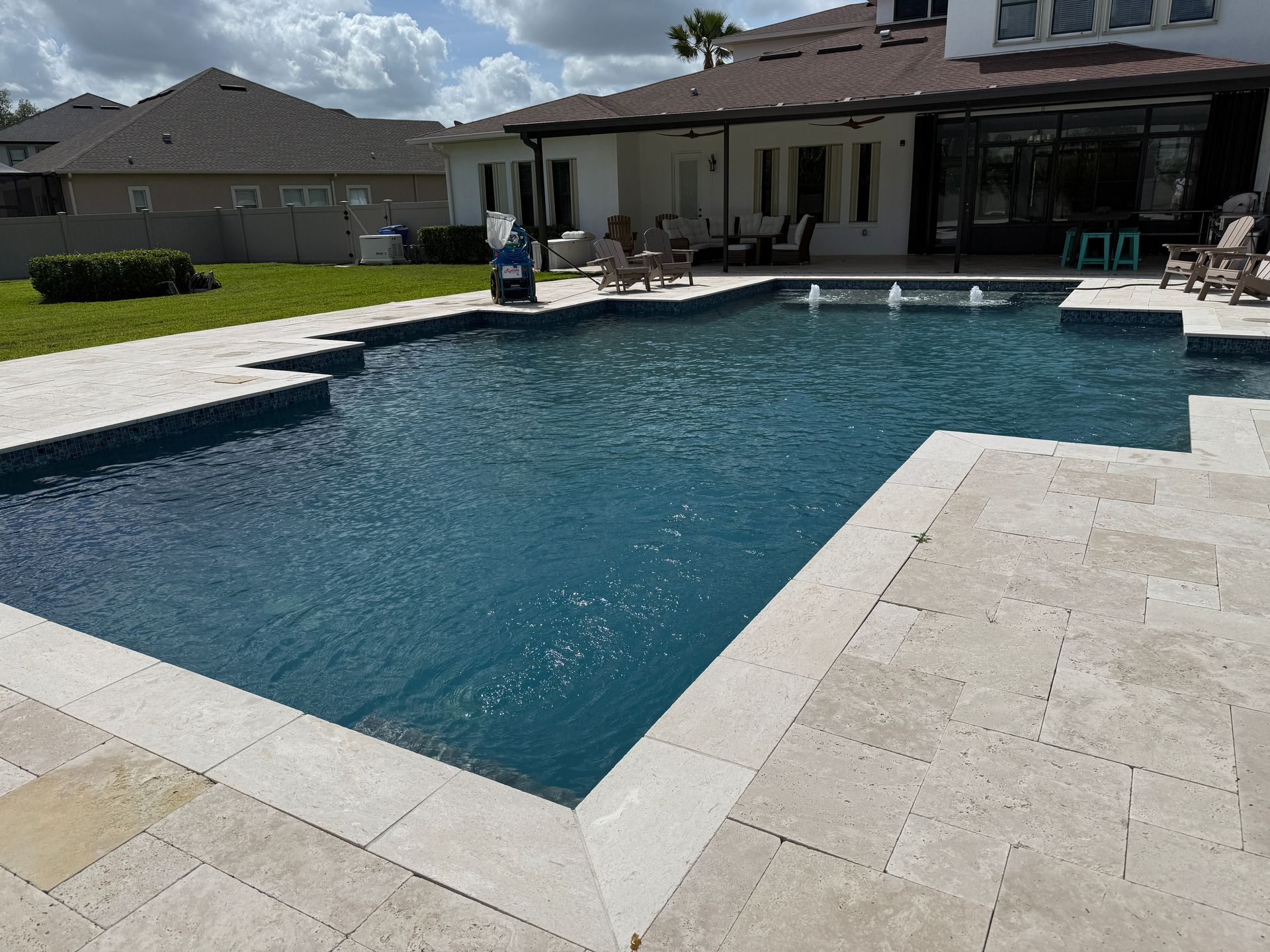 Swimming pool with travertine decking, overlooking a backyard and a two-story house.