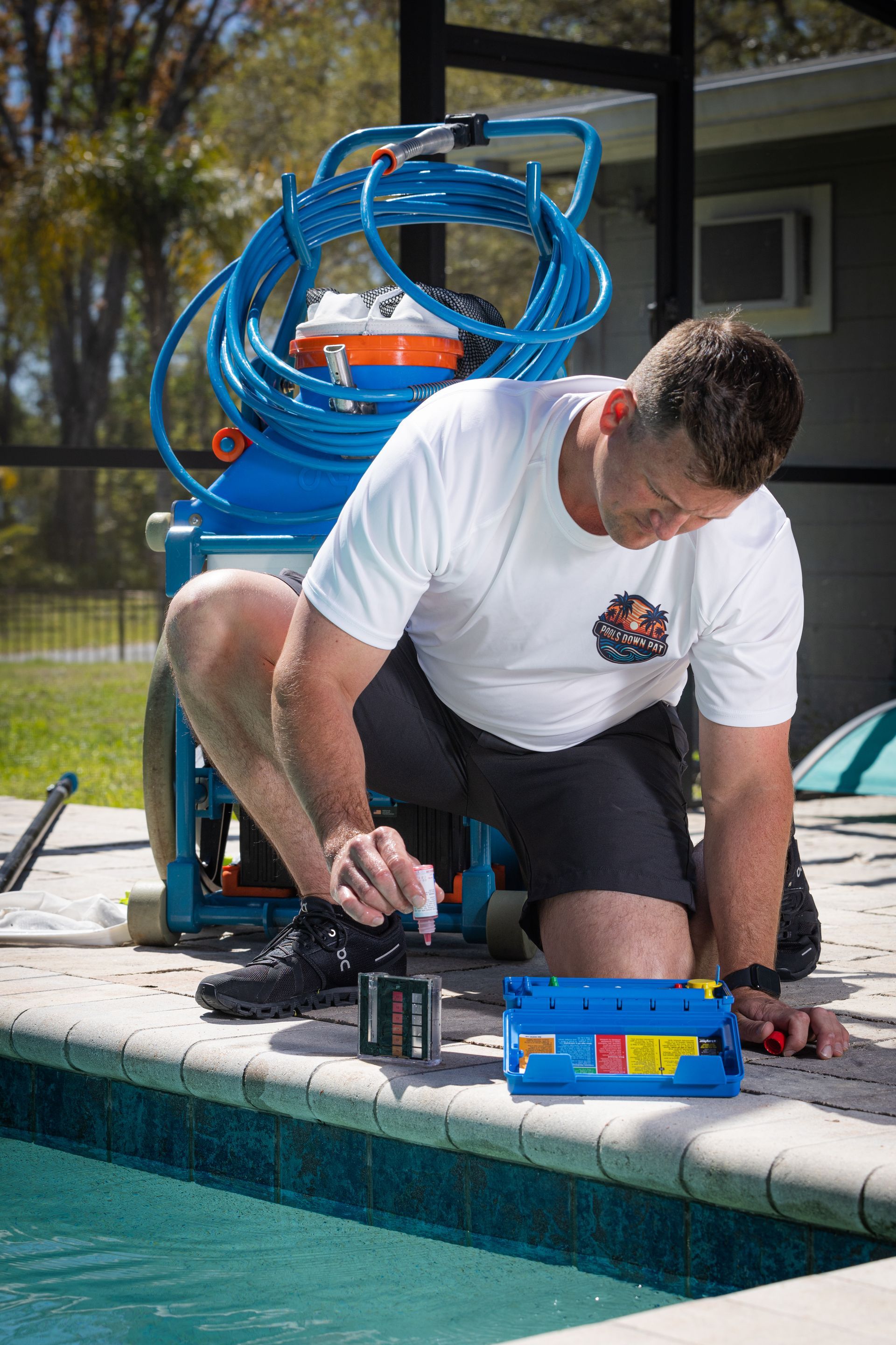 Man kneels by a pool, working on pool equipment. He wears a white shirt and black shorts. Blue equipment is behind him.