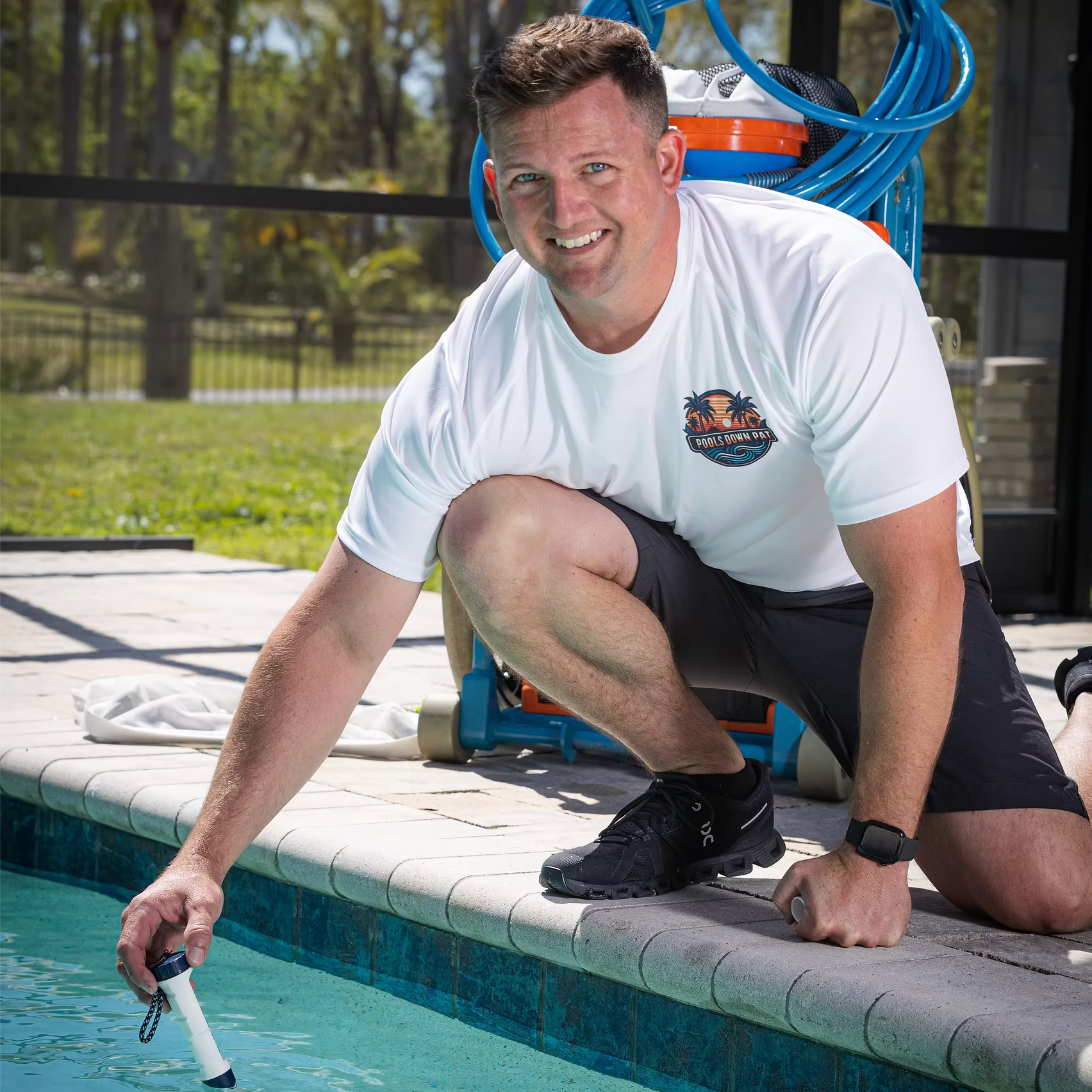 Man kneeling beside a pool, testing the water with a pool brush in a sunny backyard.
