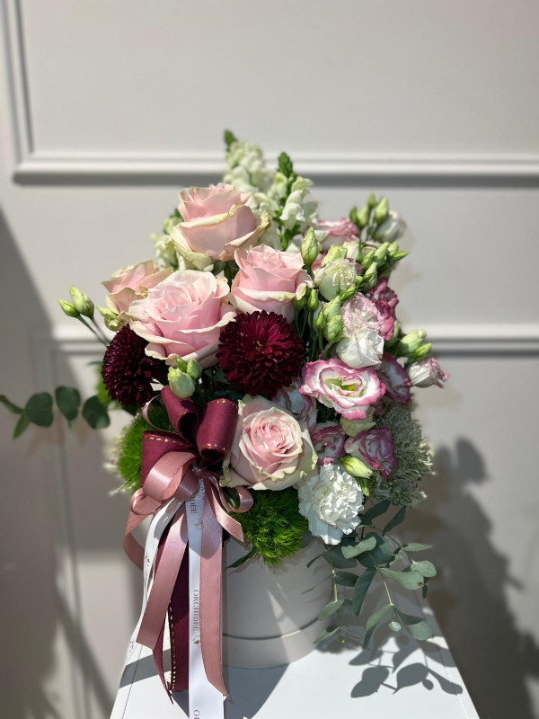 Bouquet di rose rosa e fiori color bordeaux scuro in un vaso bianco con un nastro, contro una parete chiara.