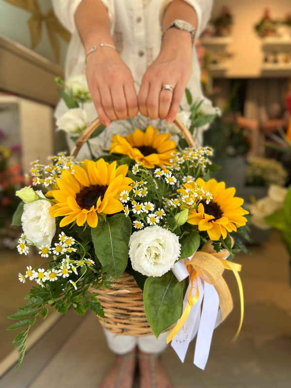 Mani che reggono un cesto di vimini con un bouquet di girasoli, rose bianche e gypsophila, legato con un nastro.