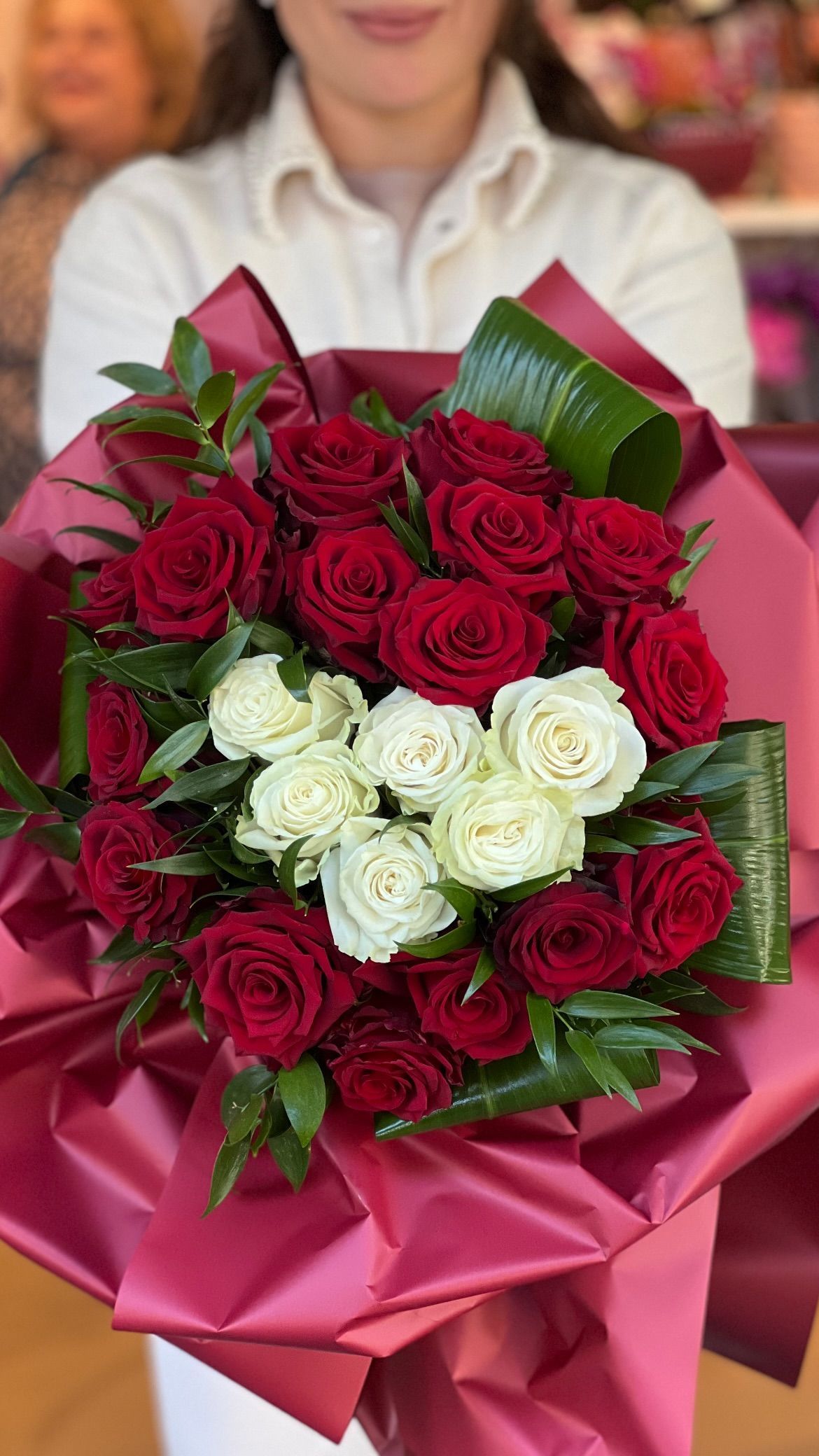 Bouquet di rose rosse e bianche avvolto in carta rosa, tenuto davanti a uno sfondo sfocato.