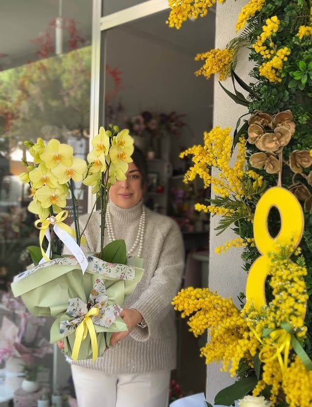 Una donna tiene in mano un mazzo di fiori giallo pallido accanto a una composizione floreale giallo brillante.