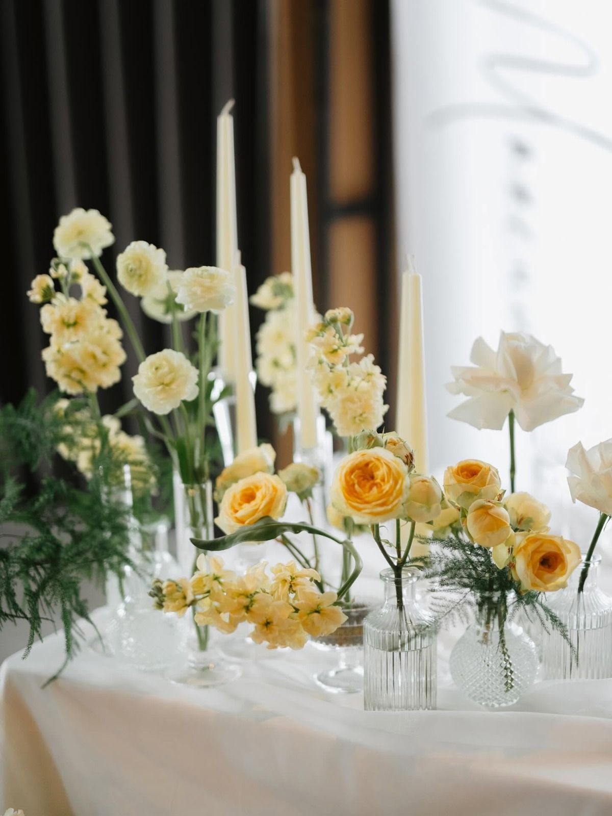 Eleganti composizioni floreali bianche e gialle con candele su un tavolo drappeggiato accanto a una finestra