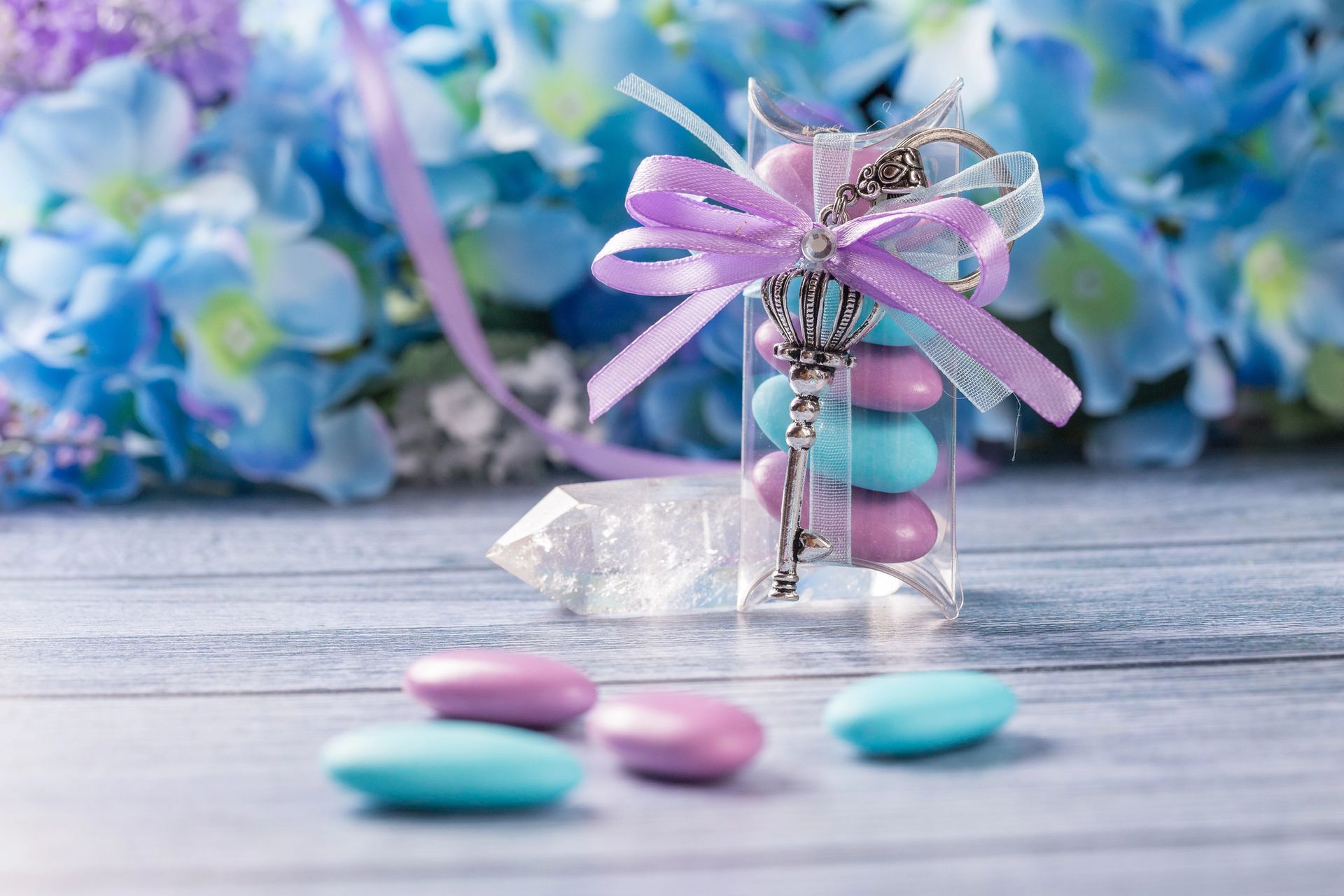 Confetti in una piccola scatola regalo con un nastro color lavanda su un tavolo, con fiori blu sullo sfondo.