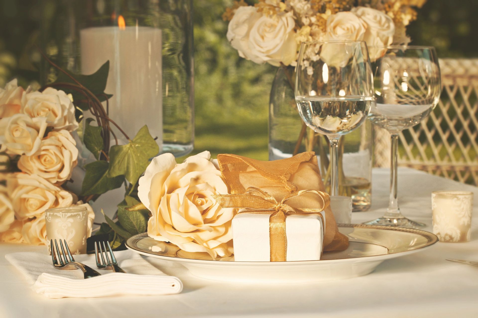 Elegante allestimento della tavola con rose color crema, calici da vino, candele e un regalo incartato all'aperto.