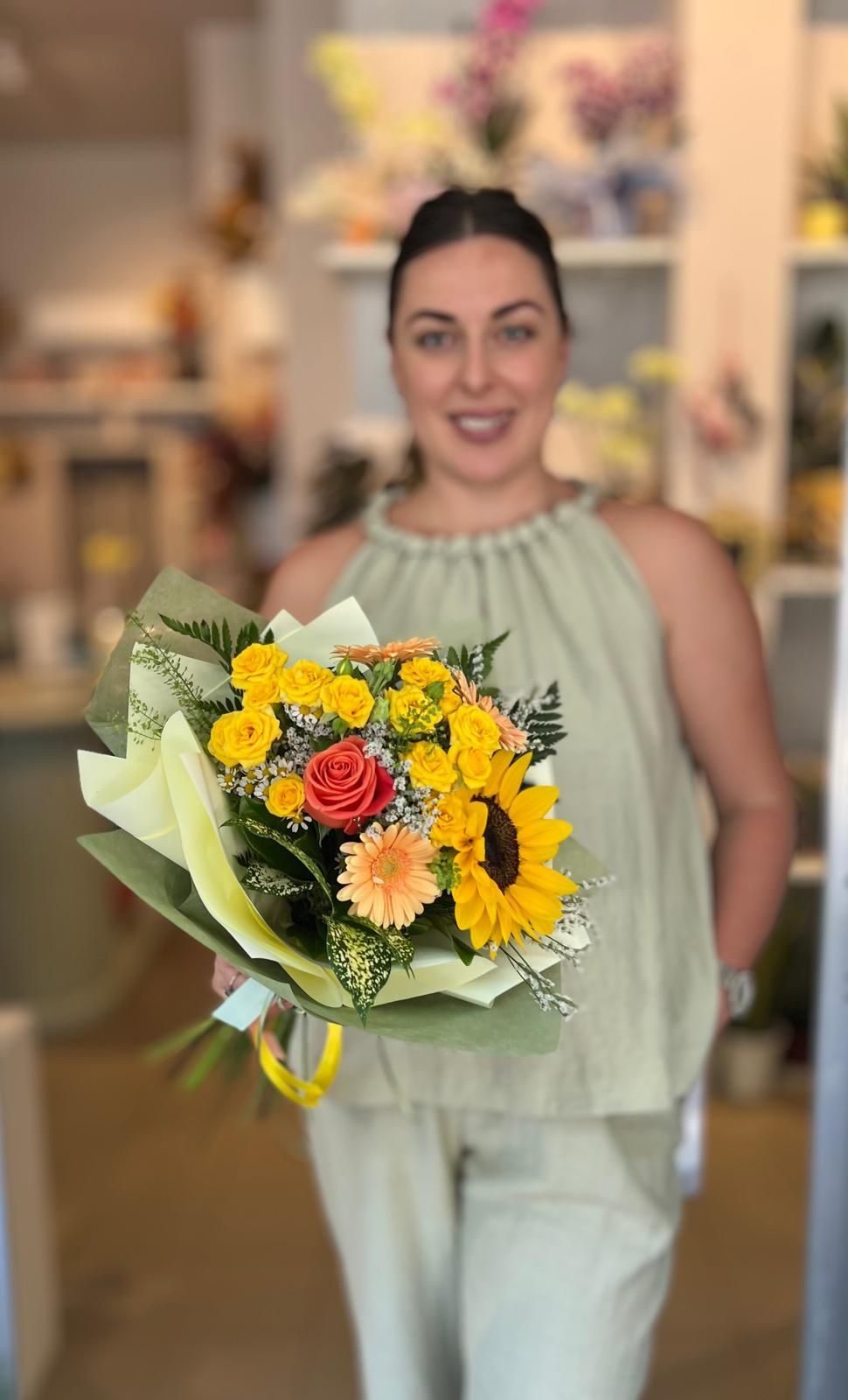 Una donna, in un ambiente interno, tiene in mano un mazzo di fiori gialli e arancioni e sorride alla telecamera.