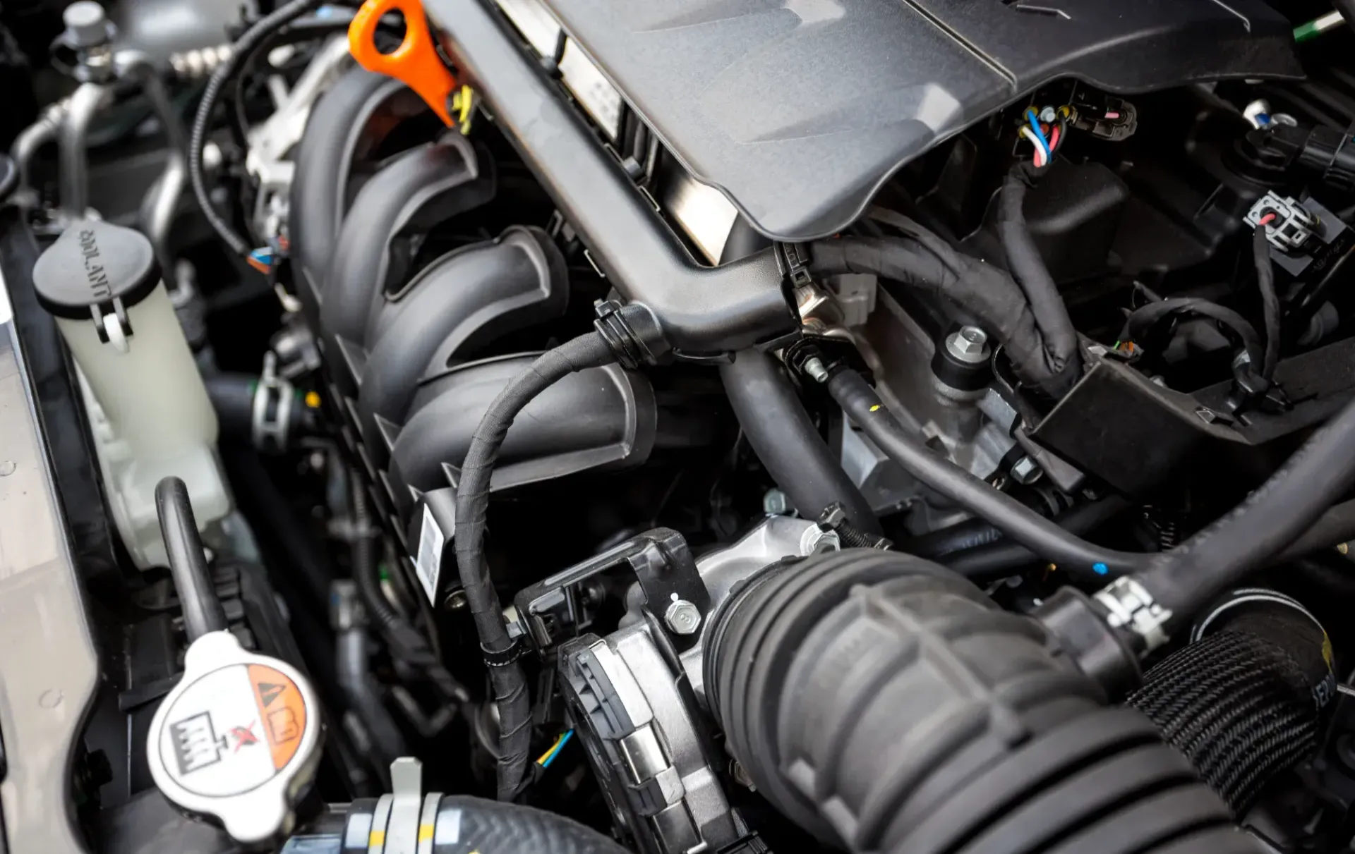 Close-up Of A Car Engine Bay With Black Intake Manifold, Hoses, And Orange Dipstick Handle