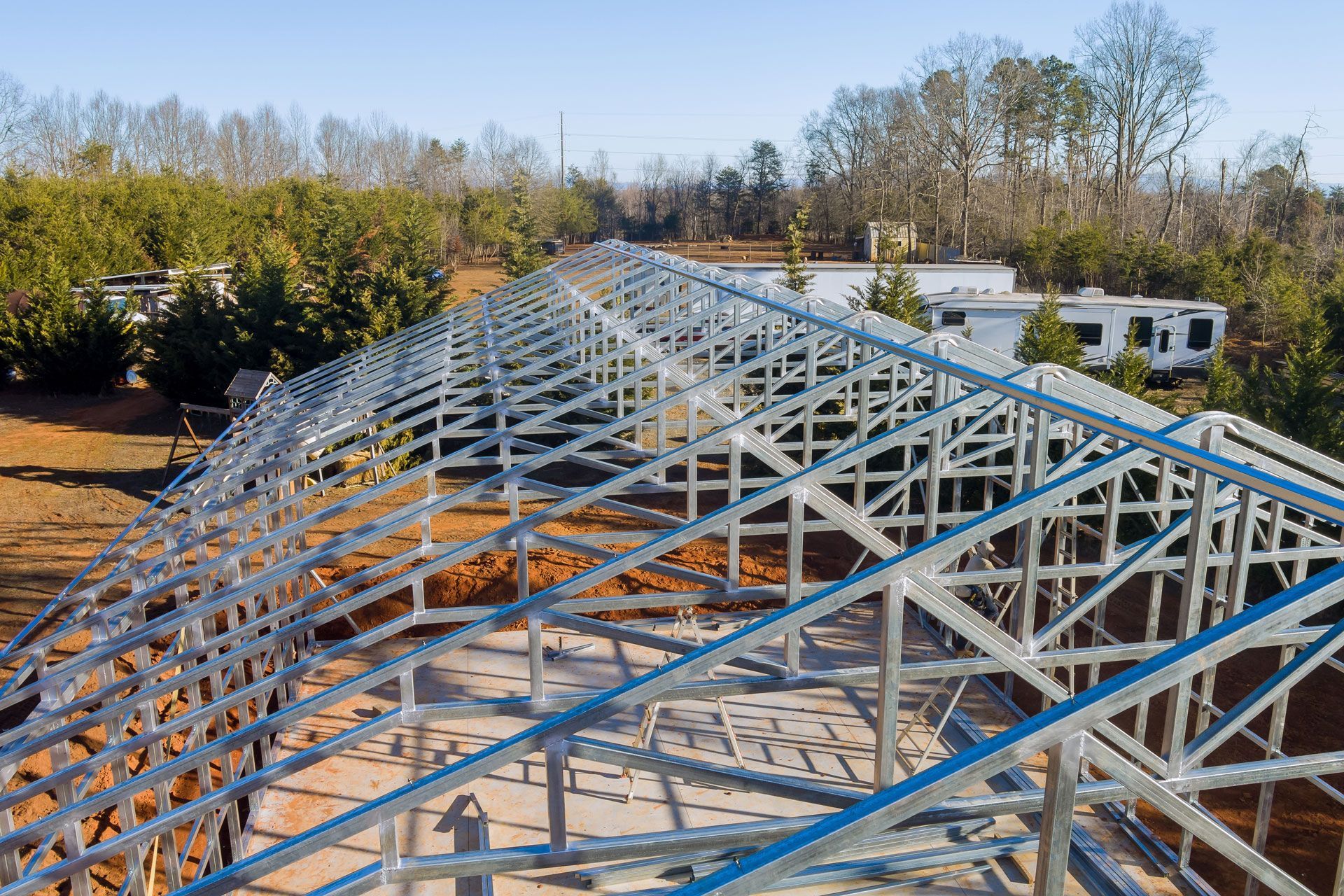 Steel roof trusses under construction on a large building site, with trees and houses in the background