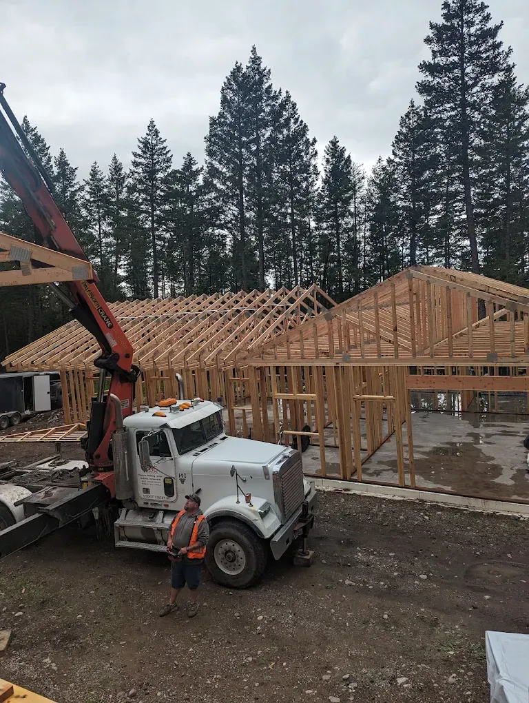 Construction site with wooden house framing, a white truck, and a worker beside an excavator in a forested area