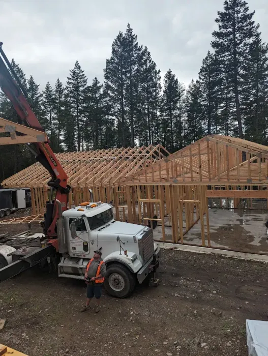 Worker beside white truck at a house framing site with an excavator and tall pine trees in the background