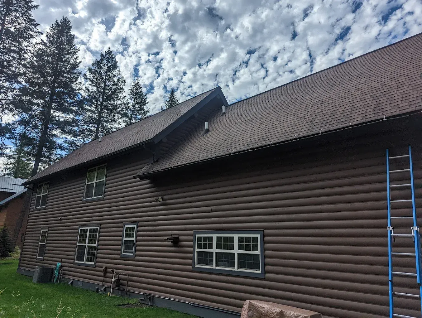 Brown log house exterior with a steep roof and blue ladder under a cloudy sky