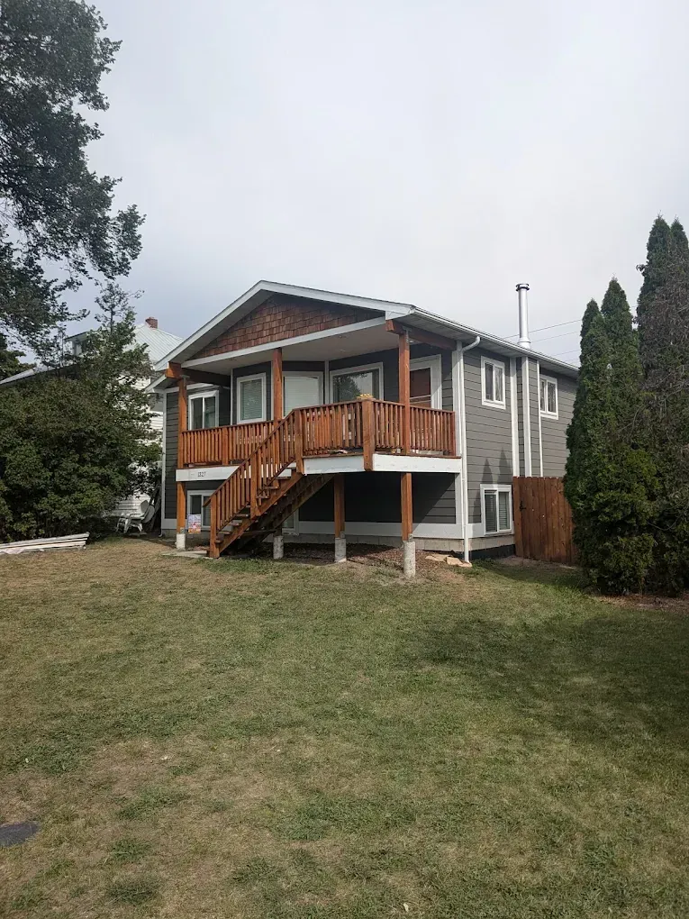 Gray house with a wooden front porch, front steps, and a large grassy yard, surrounded by trees.