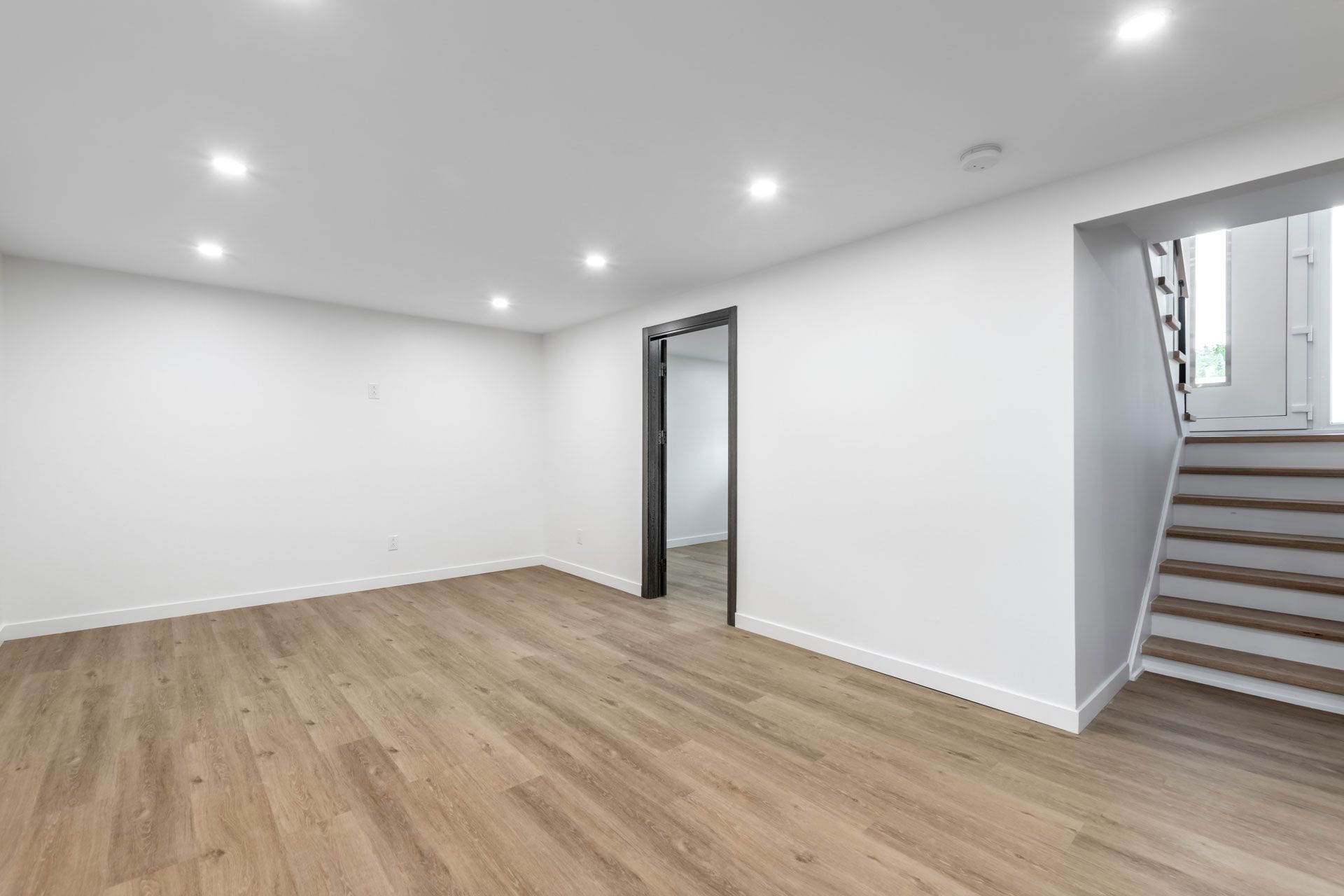 Empty modern room with light wood floors, white walls, recessed lights, and stairs beside an open doorway