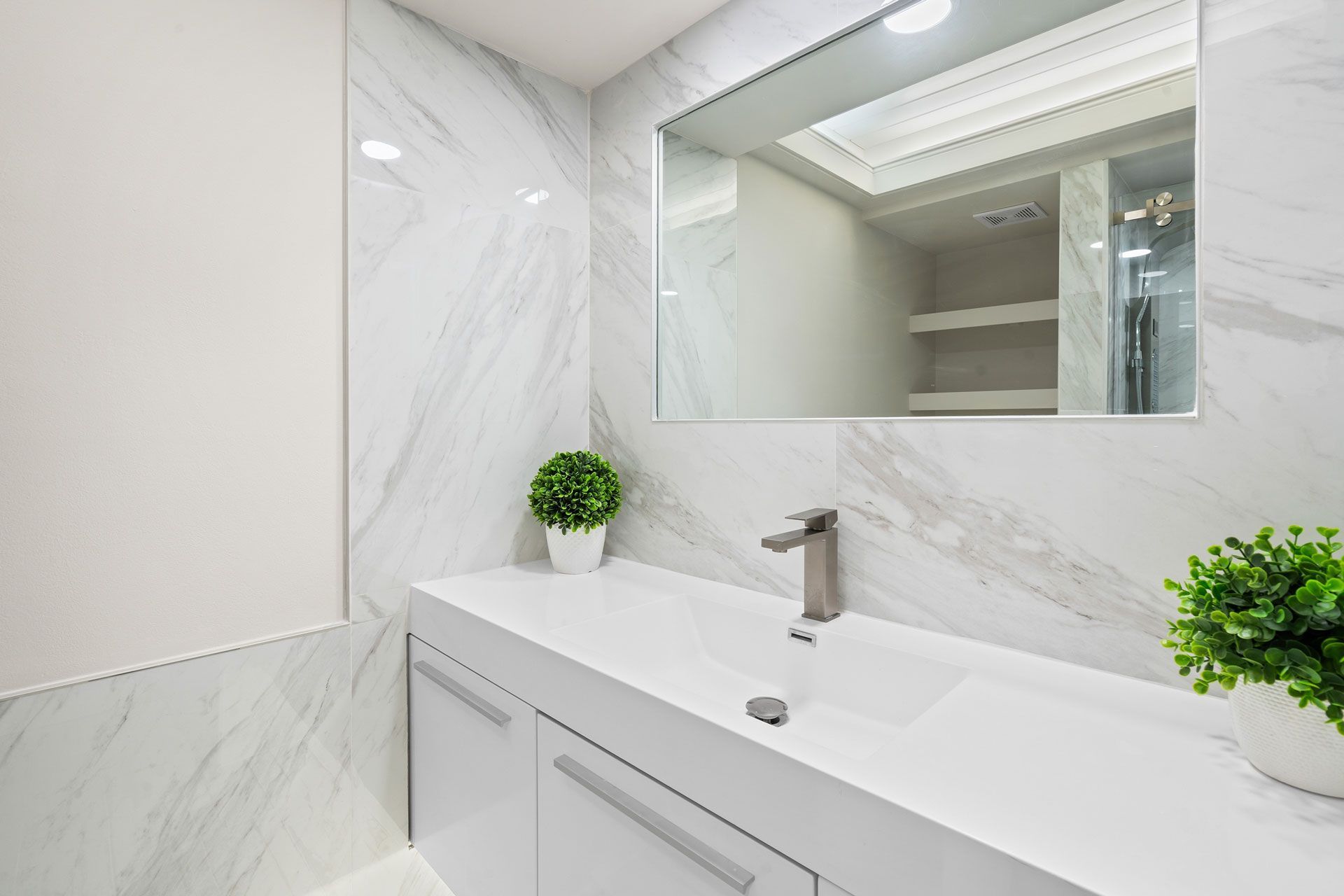 Modern white bathroom vanity with marble walls, mirror, sink, and small green plants