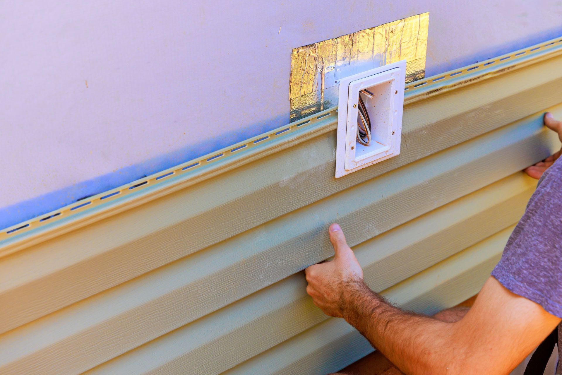 Hands installing beige siding around a white wall vent on a house exterior