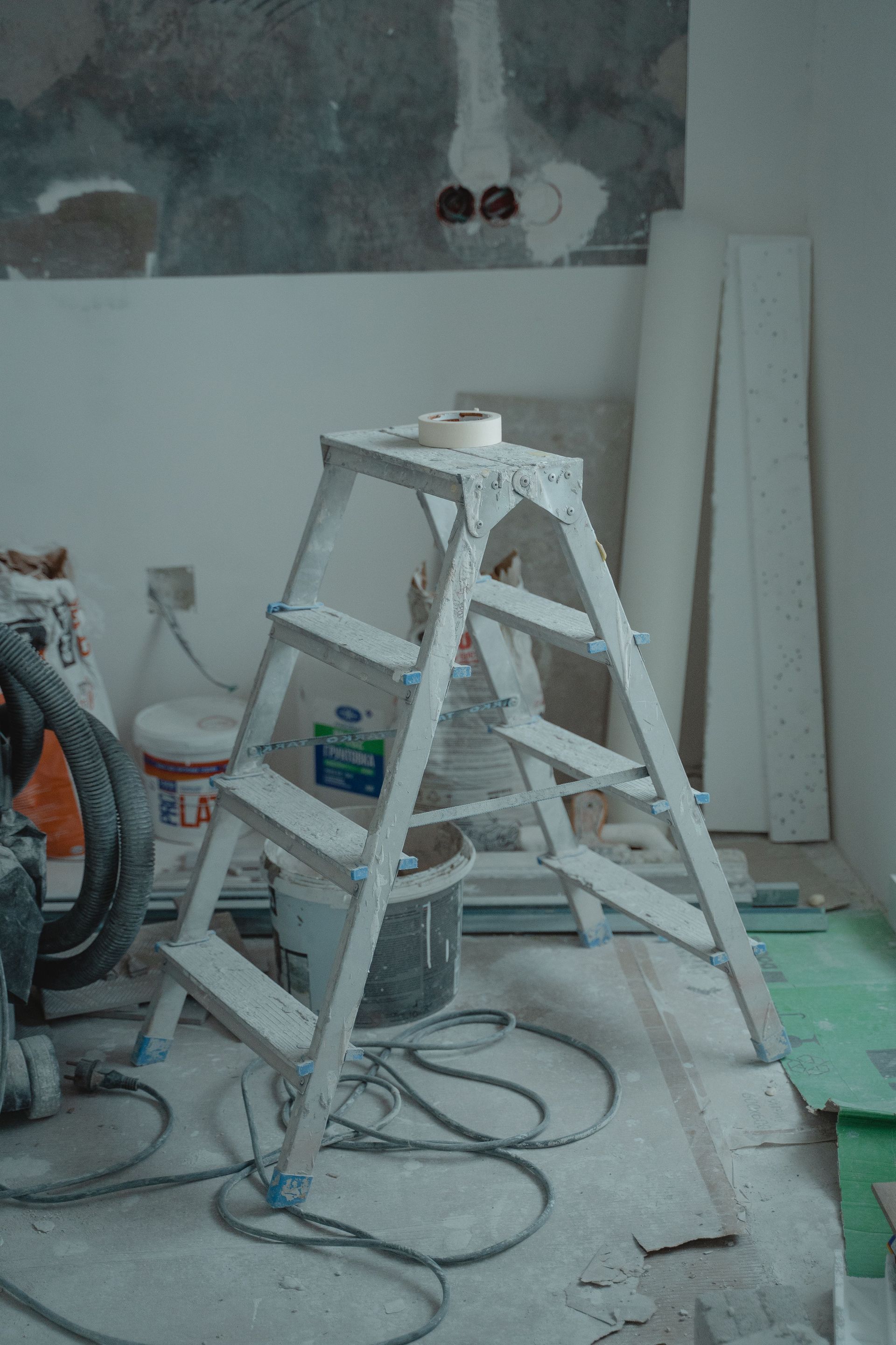White step ladder in a dusty room under renovation, with boards, cables, and tools on the floor.