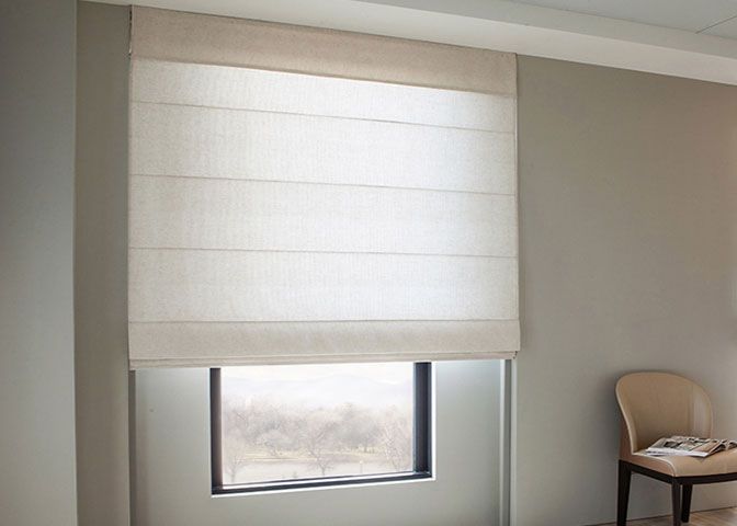 White window shade partially covering a small window beside a beige chair in a softly lit room