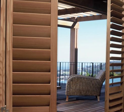 Wooden balcony with louvered shutters, a wicker chair, and ocean view beyond the railing