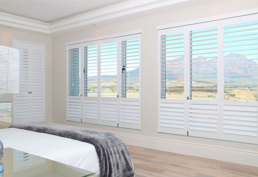Bright bedroom with white shutters, a bed, and large windows overlooking a sunny landscape