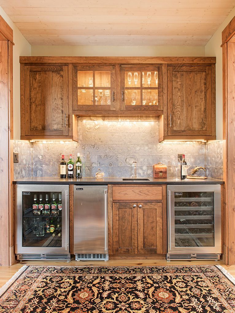 Rustic home bar with wood cabinets, stainless mini-fridges, stone backsplash, and a patterned rug
