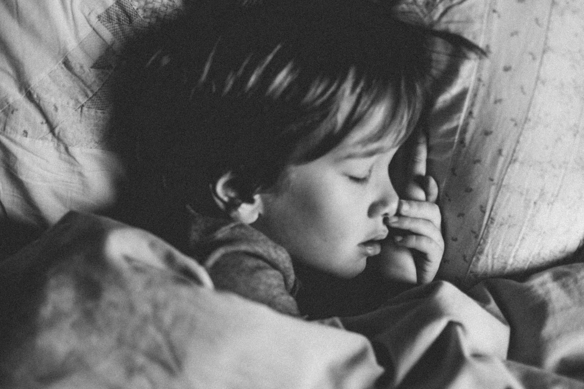Sleeping child with hand near face, tucked under a blanket on a pillow.