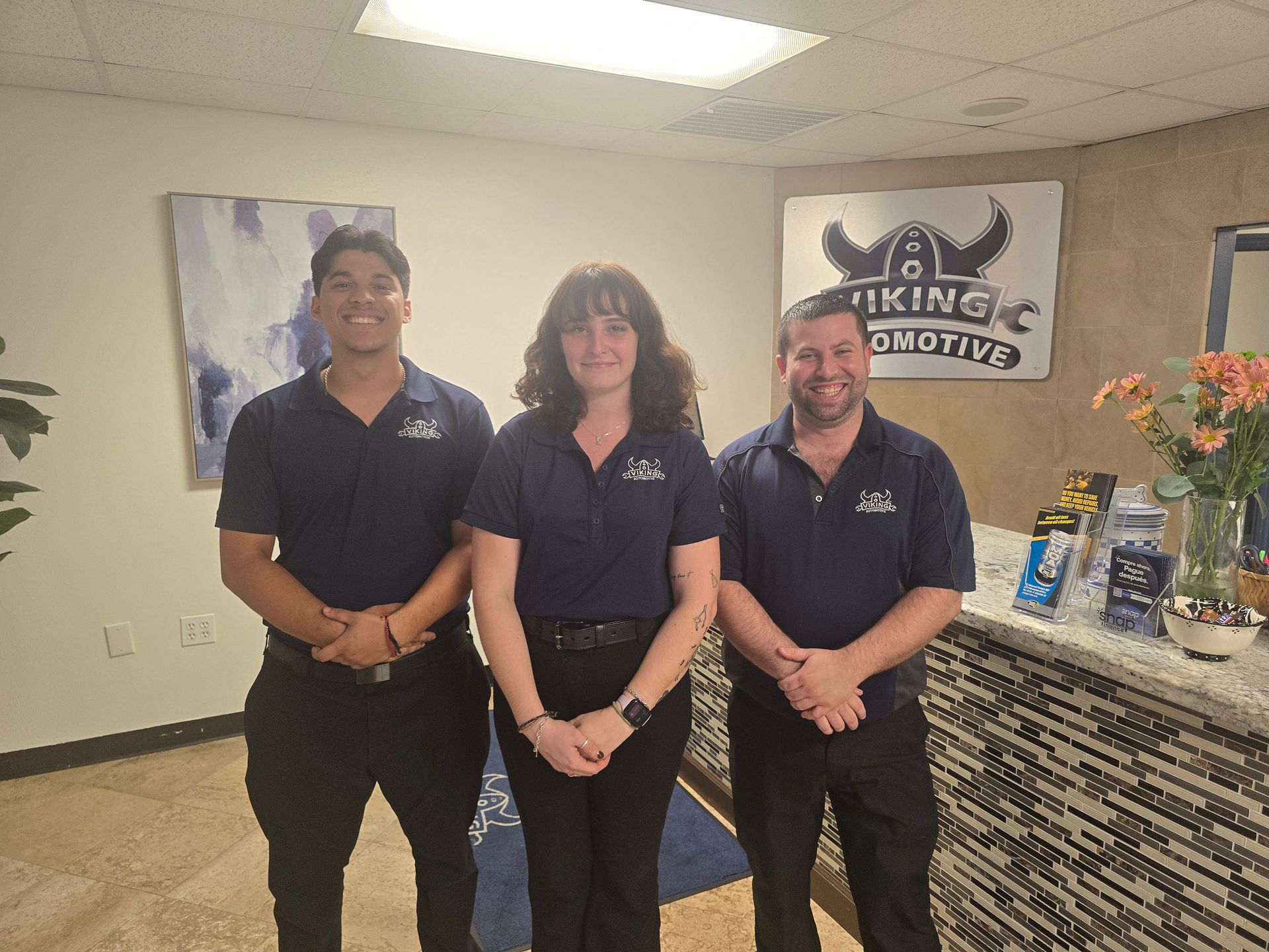 Three employees in dark uniforms stand smiling in an office lobby beside a logo sign and flowers.