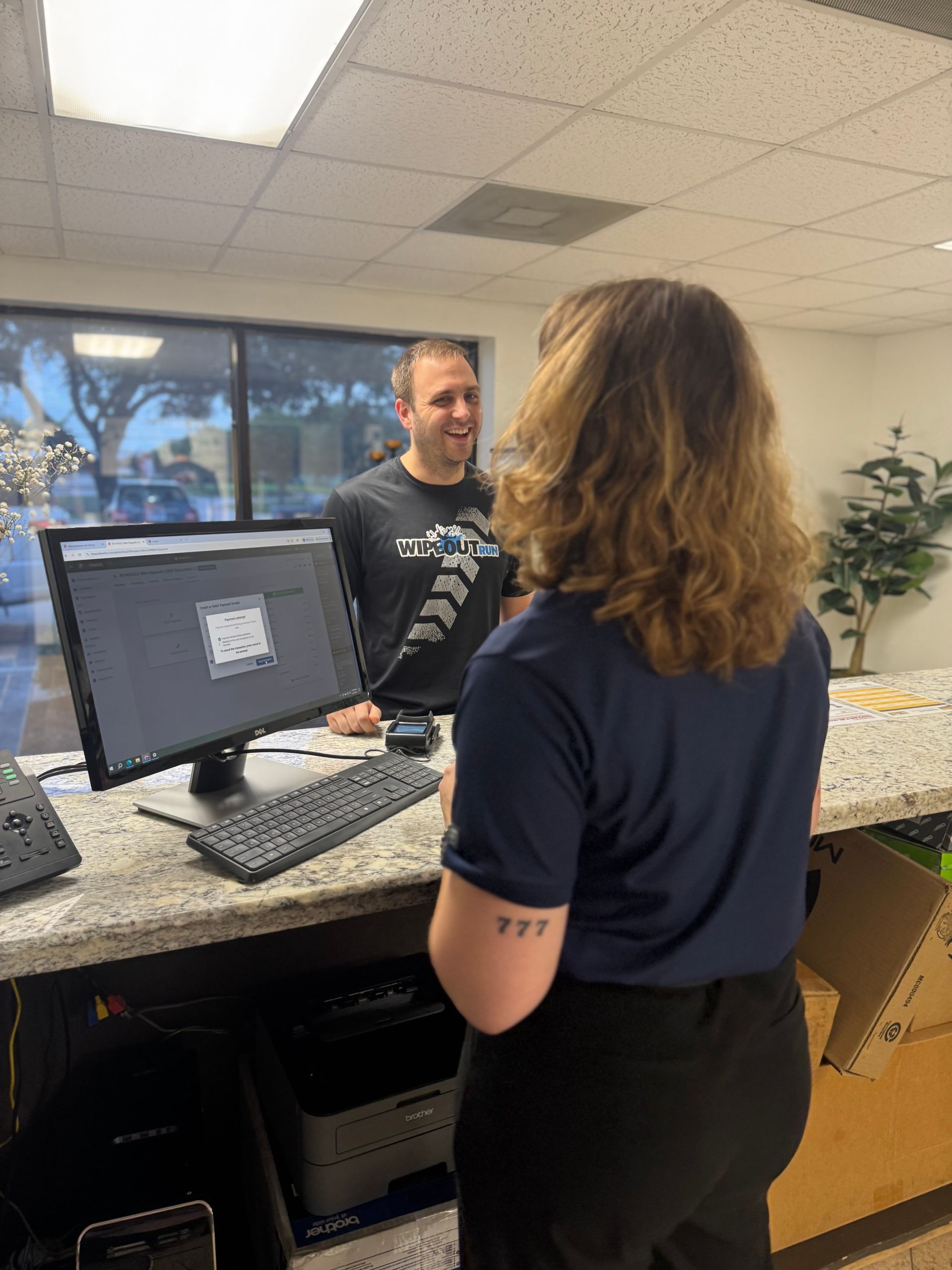 Two coworkers talking across a front desk in an office lobby, with a computer monitor and potted plant nearby.