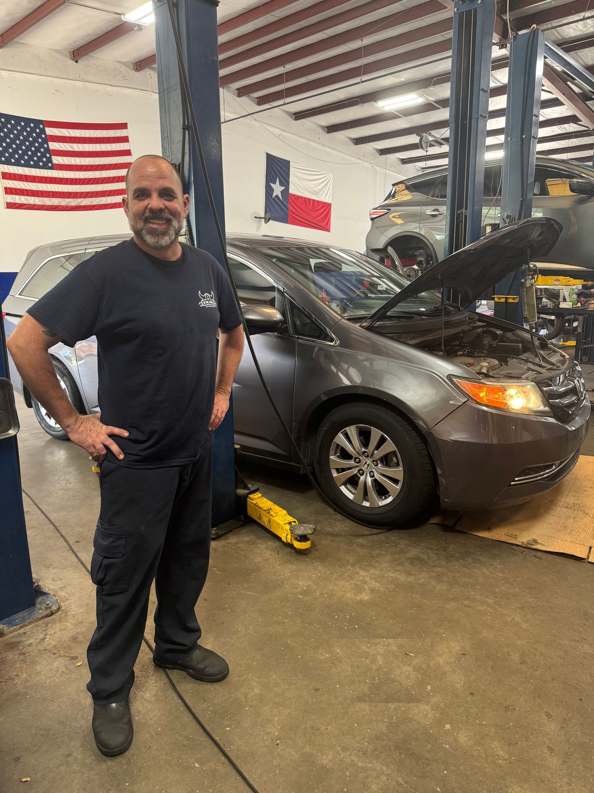 Mechanic standing beside a car with hood open in a garage, American and Texas flags in background