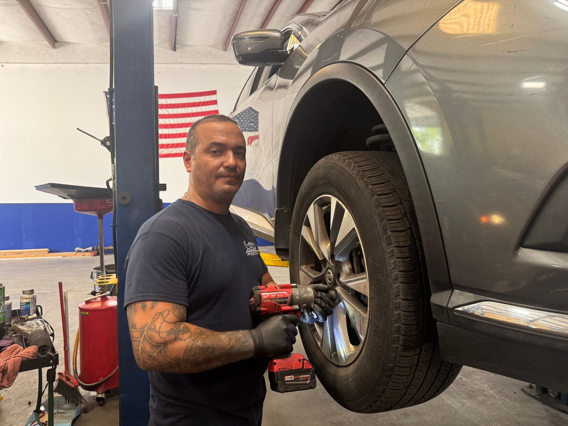 Mechanic working on a silver SUV in a garage, holding a tool near the front wheel.