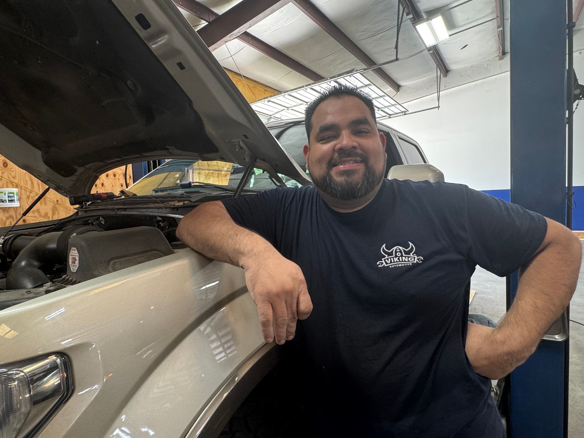 Man leaning on a beige SUV in a garage, with the hood open and workshop lights overhead.
